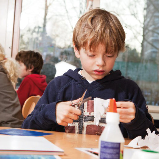 Boy in classroom cutting out paper, crafting with glue in foreground.