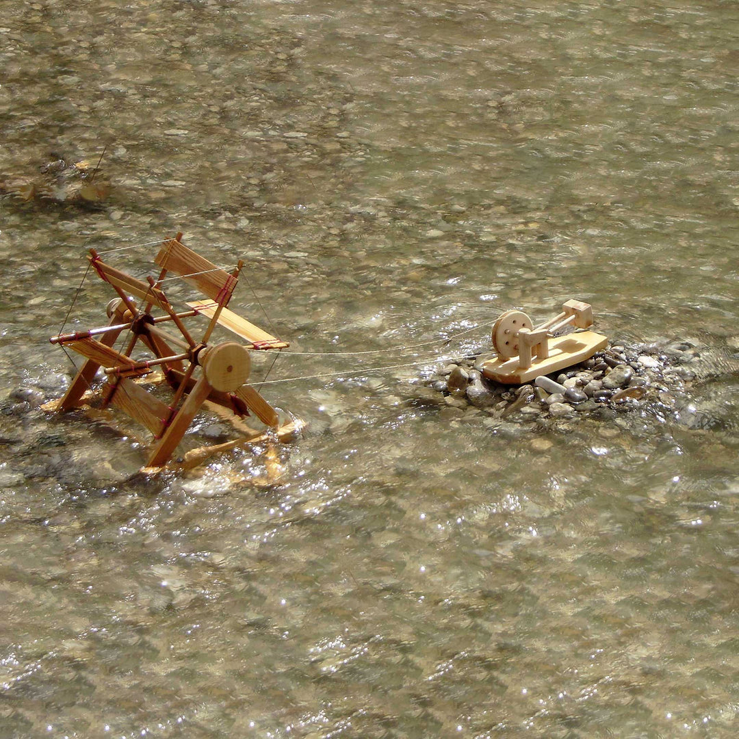 Wooden toy science models of water mill and hammer mill on a body of water with pebbles.