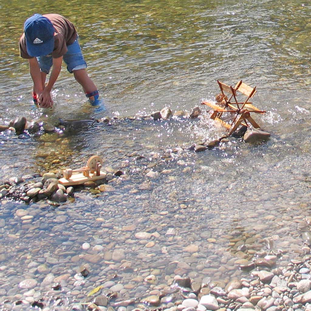 Person playing with a small wooden river wheel and hammer mill science experiments in a river with large stones, demonstrating physics principles.