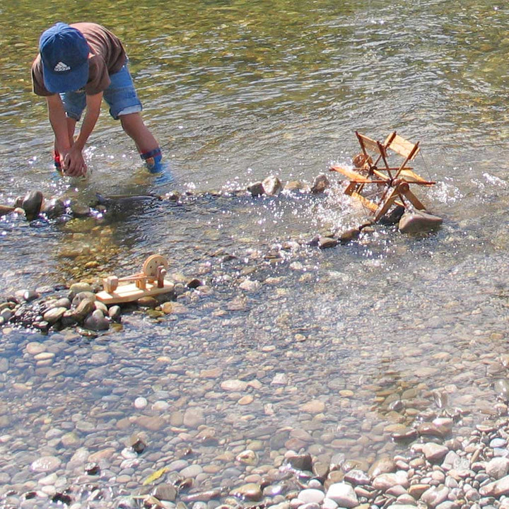 Person playing with a small wooden river wheel and hammer mill science experiments in a river with large stones, demonstrating physics principles.