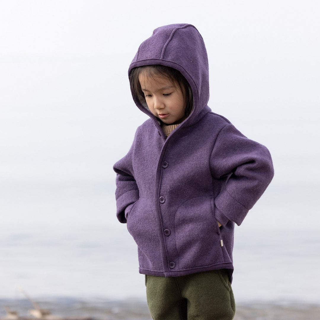 Child wearing an organic wool Disana purple coat on a beach with a cloudy sky
