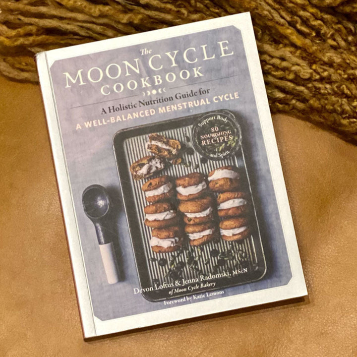 Book titled 'The Moon Cycle Cookbook' on a textured surface with a woven object in the background.