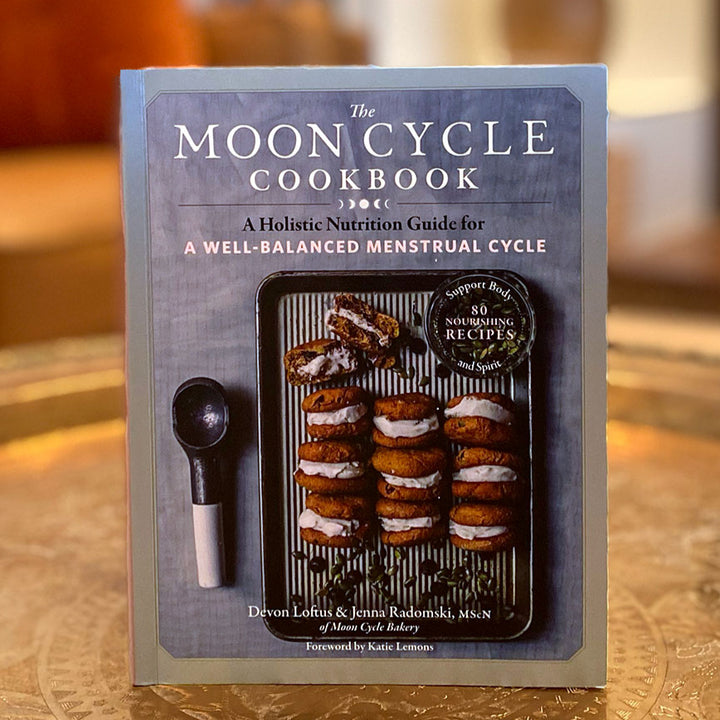 Book titled 'The Moon Cycle Cookbook' on a wooden surface with a blurred background