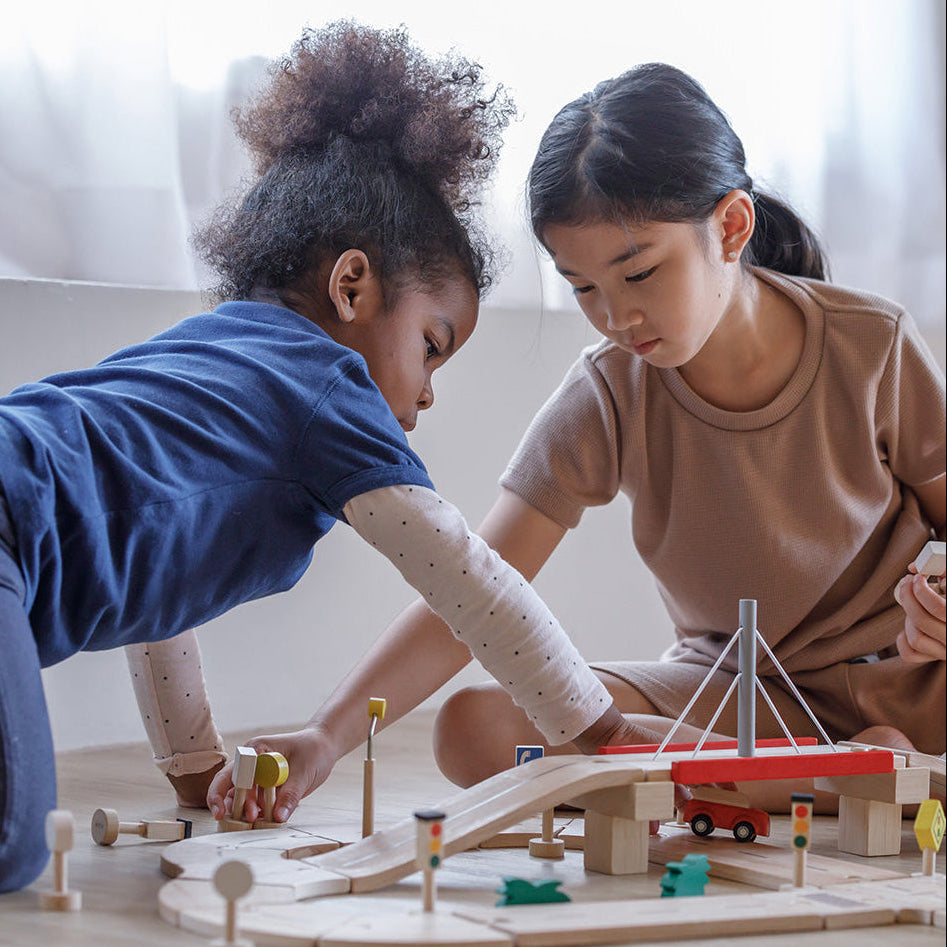 Two children playing with a wooden toy road track set on a light-colored floor.