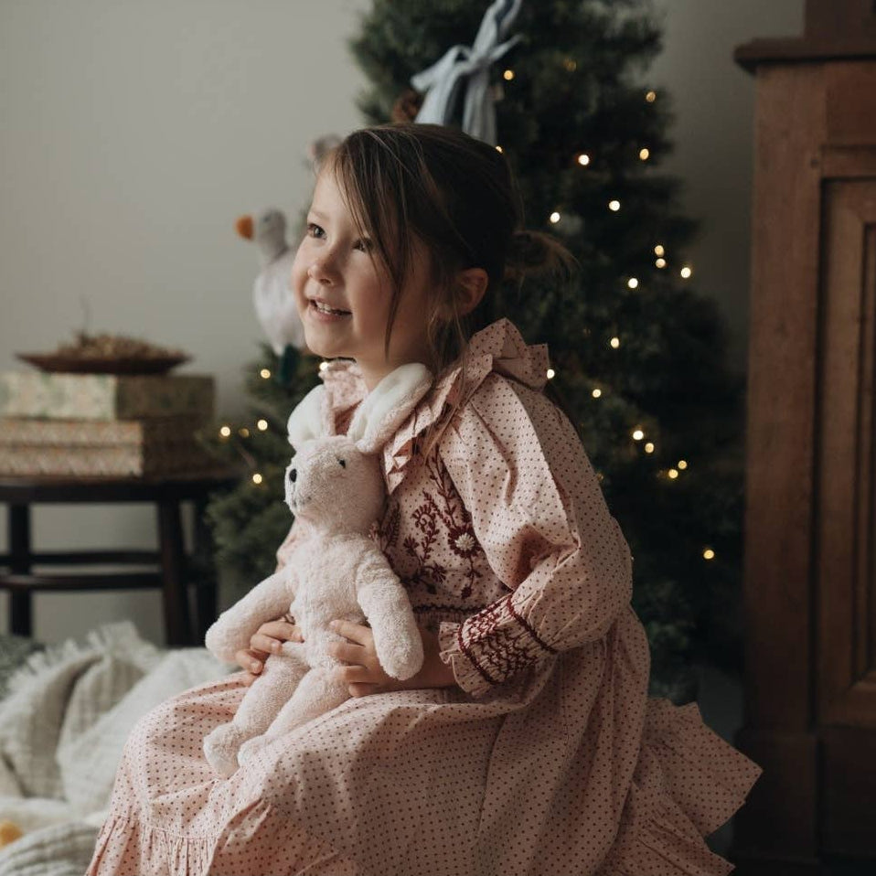Child in a pink dress holding a rose pink Senger teddy bear in a cozy room with a Christmas tree.
