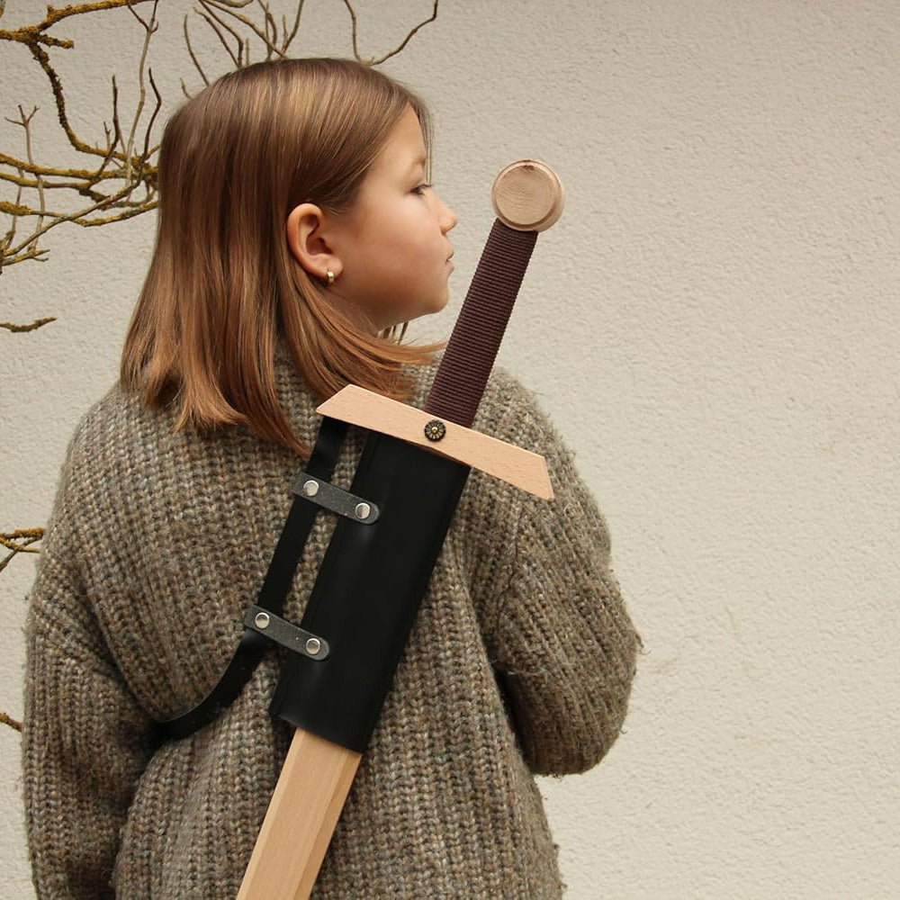 Child wearing a toy wooden sword in a holster on their back, standing against a wall.