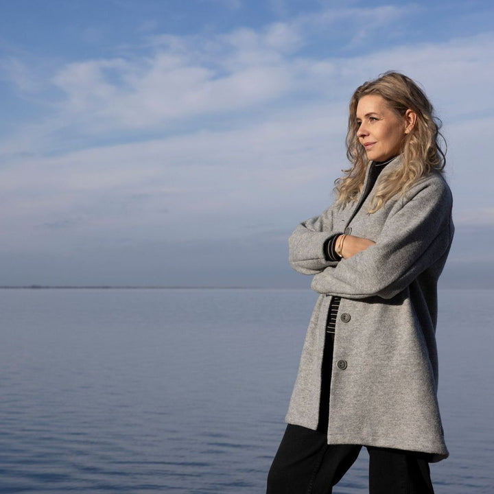 Woman in a gray organic boiled wool Disana coat standing by a body of water with a blue sky.