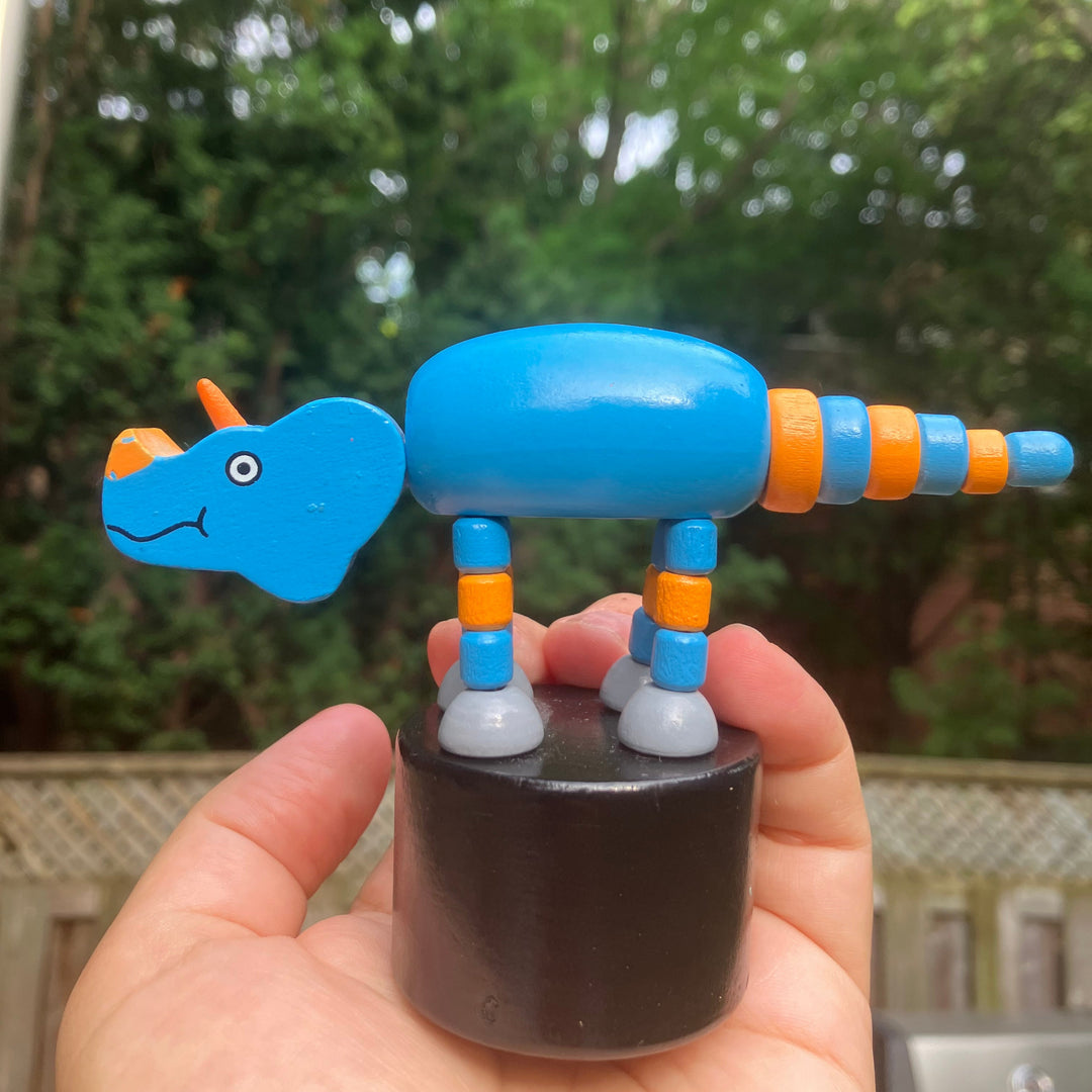 Hand holding a Jack Rabbit Creations blue and orange toy dinosaur puppet with trees in the background.