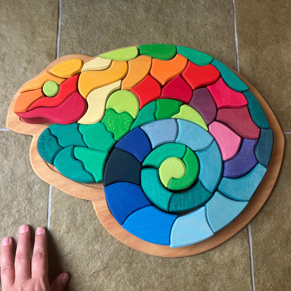 Colorful wooden puzzle shaped like a snake on a tiled floor with a hand for scale.