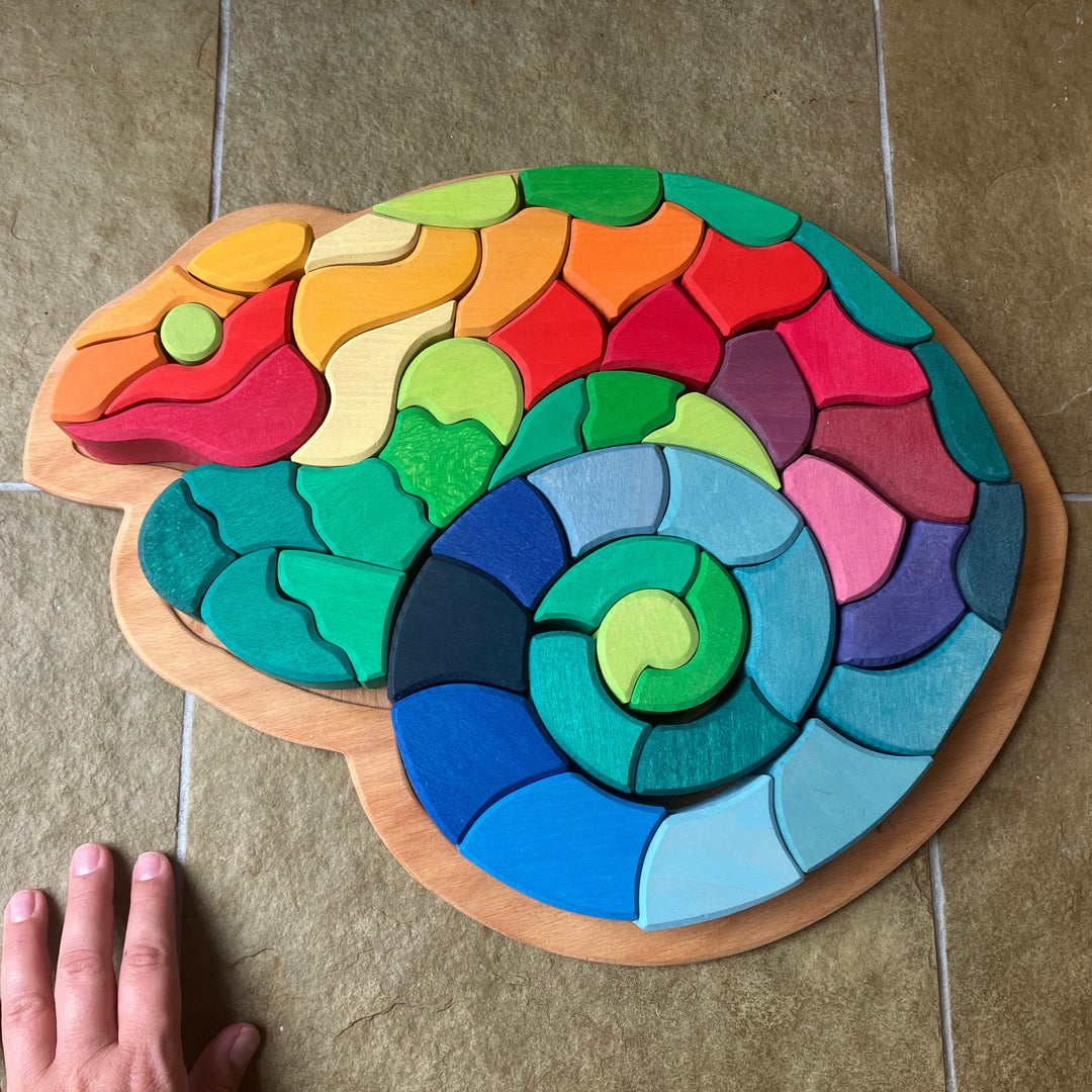 Colorful wooden puzzle shaped like a snake on a tiled floor with a hand for scale.