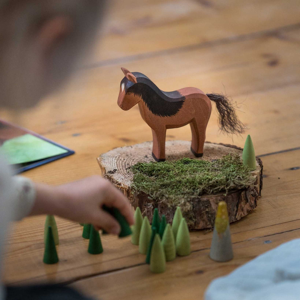 A child's hand playing with Ostheimer Hrókur wooden toy horse on tree trunk platter with moss and Grapat wooden green cones and trees on the floor. 