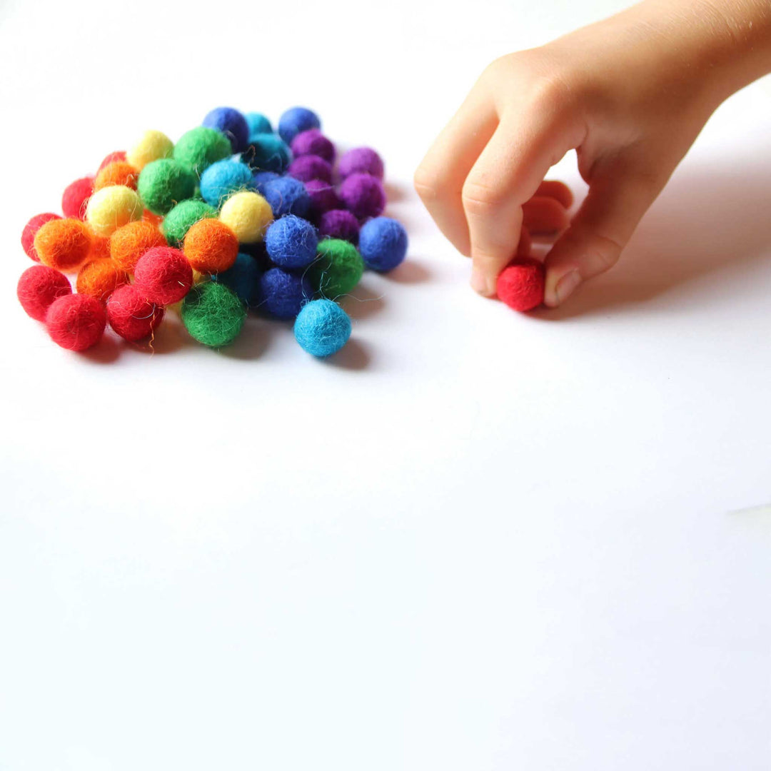 Colourful mix of 1/2 inch wool balls, with child's hand holding a red ball pinched between forefinger and thumb on a white surface.