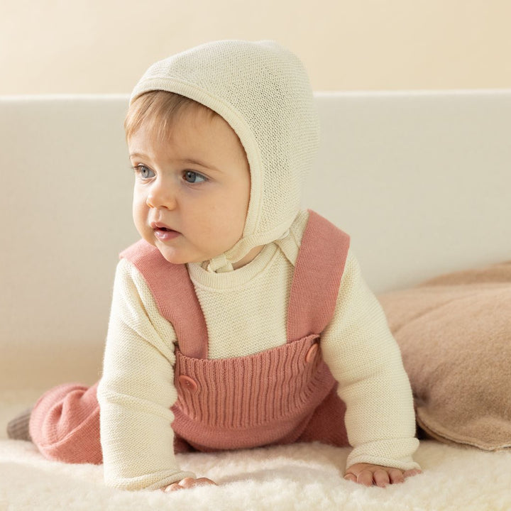 Baby on plush wool carpet, with Disana organic merino wool natural white knitted bonnet, white knit sweater, and pink rompers.
