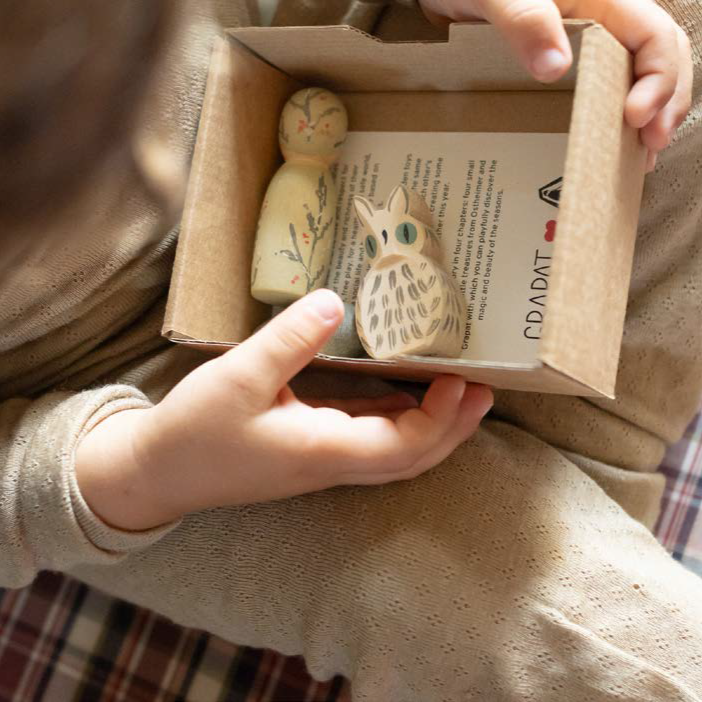 Child holding a small cardboard box with Ostheimer mini owl and Grapat winter peg doll wooden figurines and text inside.