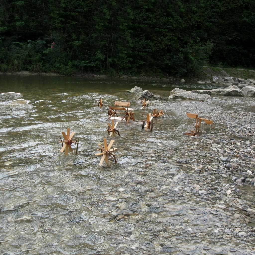 River wheel kit with small waterwheels and water see-saw, set up in shallow water by a riverbank with pebbles and coniferous trees in the background