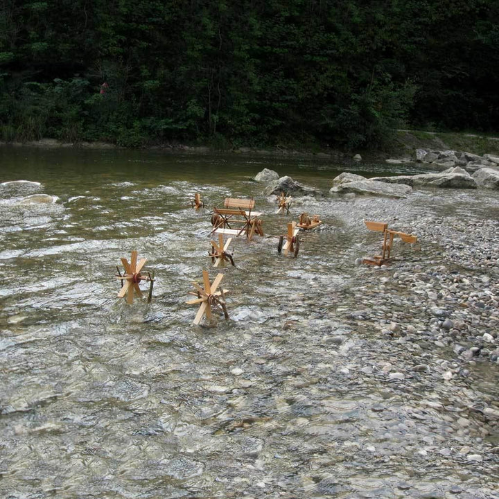 River wheel kit with small waterwheels and water see-saw, set up in shallow water by a riverbank with pebbles and coniferous trees in the background